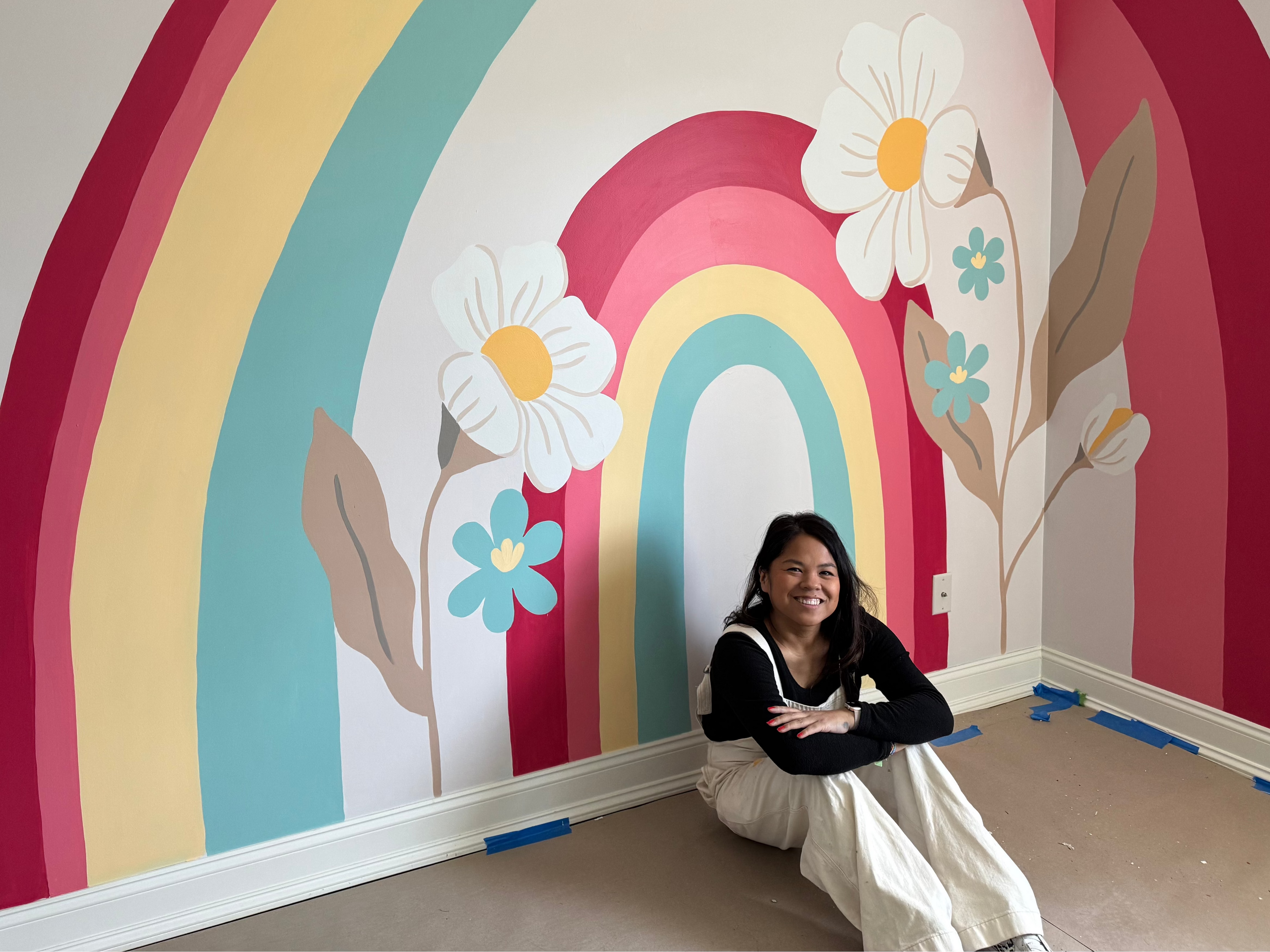 Woman sitting in front of a colorful rainbow and flower mural on a wall.